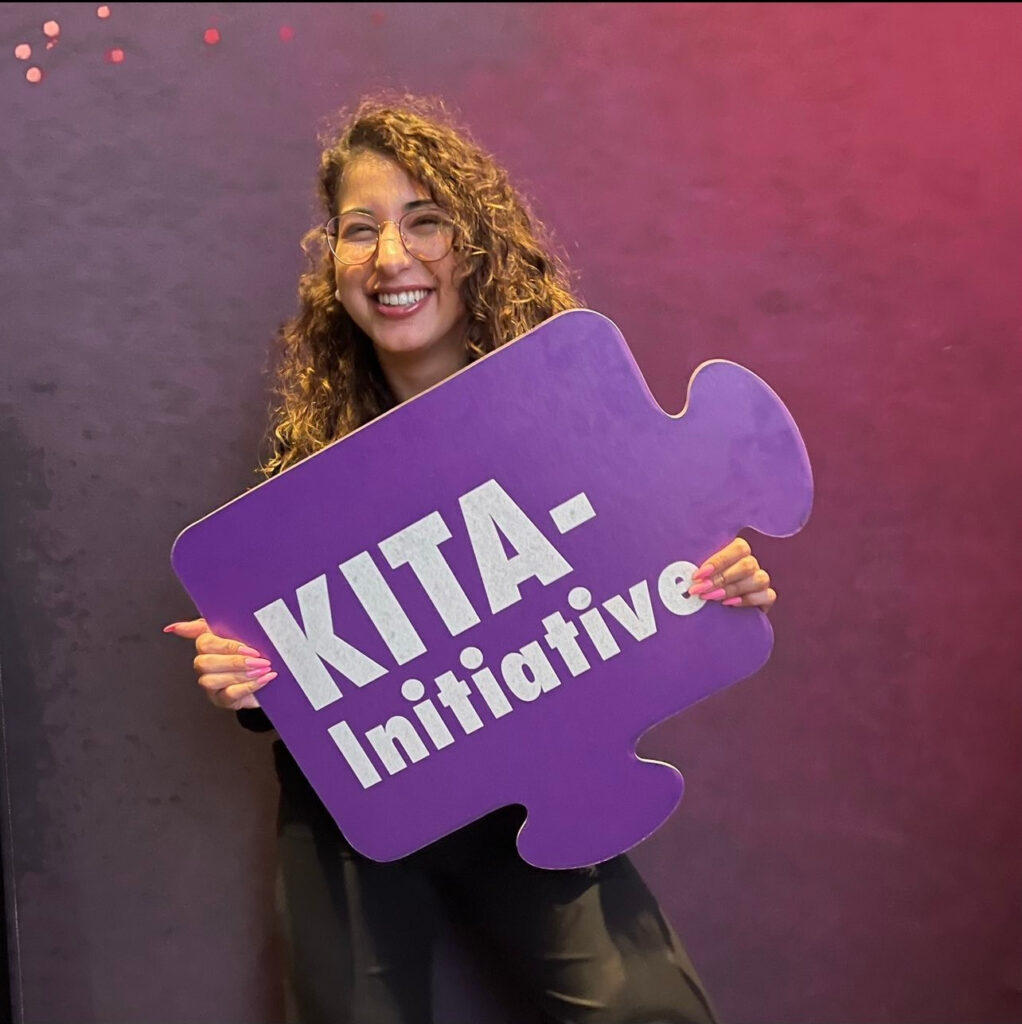 Maria lächelt und hält ein Puzzle-Stück aus einer Werbung für die KITA-Initiative in der Hand.